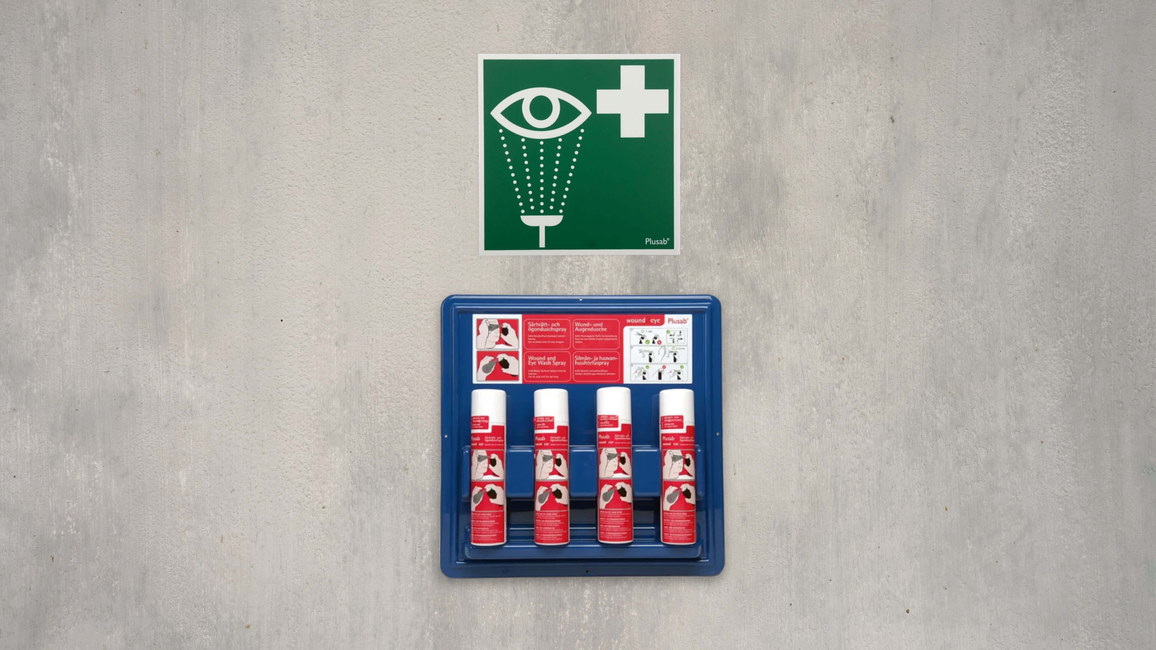 En blå hållare med fyra flaskor ögondusch sitter på en betongvägg. Ovanför sitter en grön skylt som indikerar ögondusch.